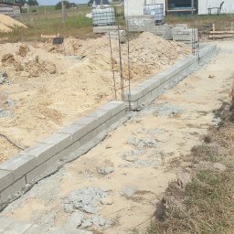 Bartłomiej Sokołowski - Budowa fundamentów: wzniesiony murek z bloczków betonowych, wystające pręty zbrojeniowe, dom w tle, słoneczny dzień, plac budowy w trakcie realizacji.
