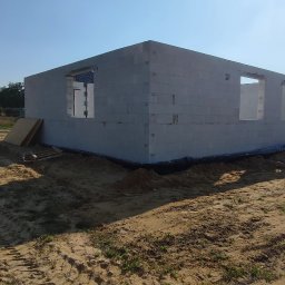 Bartłomiej Sokołowski - Surowy stan: ściany z betonu komórkowego z otworami okiennymi wzniesione na piaszczystym gruncie, w tle błękitne niebo i zieleń. Widoczne deski i ślady opon.