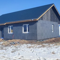 Bartłomiej Sokołowski - Niedokończony dom z szarej kostki, z oknami i ciemnym dachem, w zimowym krajobrazie z resztkami śniegu na ziemi i błękitnym niebem.