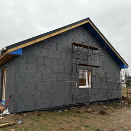 Bartłomiej Sokołowski - Budowa domu: ściany zewnętrzne z szarej izolacji, rusztowanie przy oknie. Widoczne drewniane elementy konstrukcji dachu i fragment podwórka z resztkami materiałów.