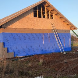 Bartłomiej Sokołowski - Budowa domu szkieletowego: ściany z izolacją, widoczna konstrukcja dachu z oknem. Drabina oparta o ścianę, teren budowy z roślinnością.