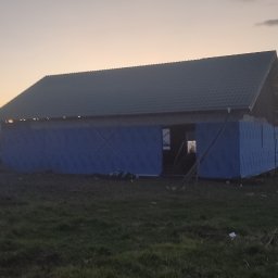 Bartłomiej Sokołowski - Wznoszony dom z ciemnym dachem i niebieską izolacją na tle zachodzącego słońca. Widoczne zarysy okien i drzwi. Prace budowlane w toku.