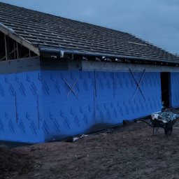 Bartłomiej Sokołowski - Budowa domu: ściana z niebieską izolacją, widoczny szkielet dachu i taczka. Etap prac budowlanych przy wznoszeniu domu, wieczorne oświetlenie.