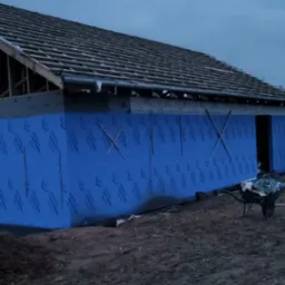 Budowa domu: ściana z niebieską izolacją, widoczny szkielet dachu i taczka. Etap prac budowlanych przy wznoszeniu domu, wieczorne oświetlenie.