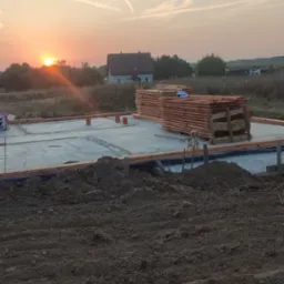 Fundamenty domu jednorodzinnego o zachodzie słońca. Na pierwszym planie wykop, w tle drewno budowlane i zarys domu. Widoczny pies rasy shih tzu.