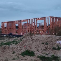 Bartłomiej Sokołowski - Konstrukcja szkieletowa domu w trakcie budowy, widok na drewniany szkielet z otworami okiennymi na tle pochmurnego nieba i piaszczystego terenu.