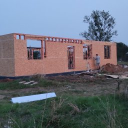 Bartłomiej Sokołowski - Wznoszenie ścian domu szkieletowego z płyt OSB, widoczny fragment konstrukcji i pracujący mężczyzna. W tle zachodzące słońce nadaje zdjęciu ciepły koloryt.