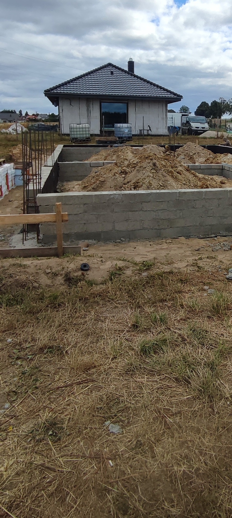 Wznoszenie fundamentów domu jednorodzinnego: betonowe ściany szalunkowe, zbrojenie pionowe. W tle budynek w stanie surowym zamkniętym i zaparkowany samochód dostawczy.