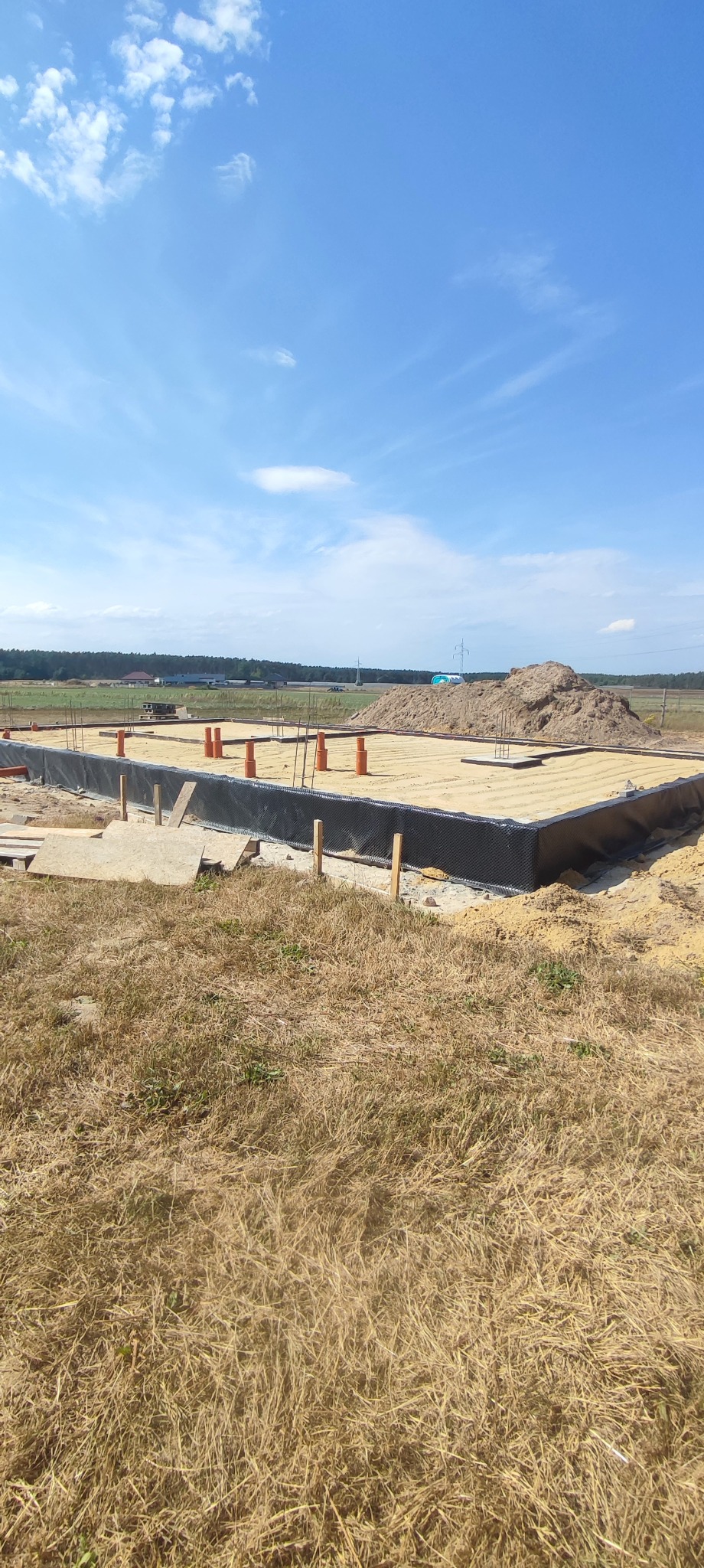 Przygotowany fundament pod budowę domu, widoczne rury, zbrojenie i folia izolacyjna. W tle krajobraz z lasem i błękitnym niebem.