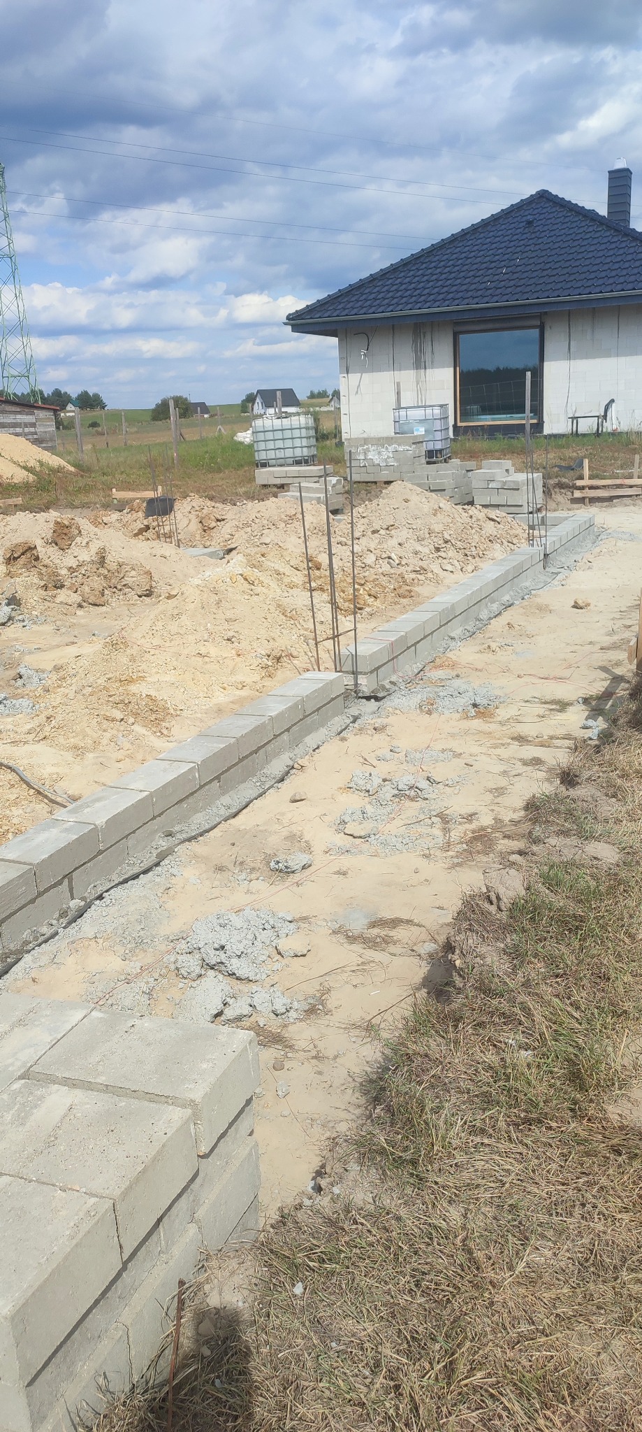 Budowa fundamentów: wzniesiony murek z bloczków betonowych, wystające pręty zbrojeniowe, dom w tle, słoneczny dzień, plac budowy w trakcie realizacji.