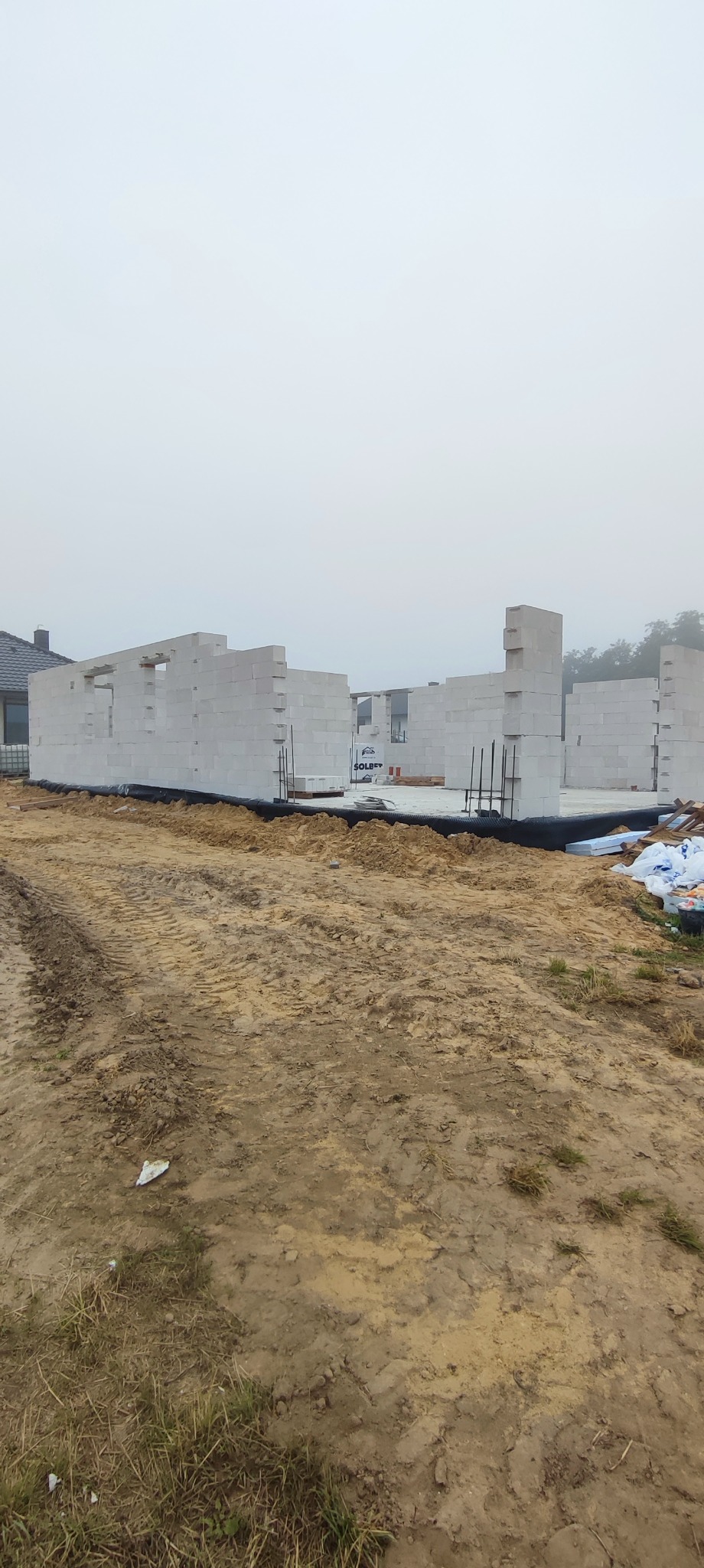Budowa domu w trakcie: wzniesione ściany z betonu komórkowego na fundamencie, widoczne zbrojenia i materiały budowlane. Mglista pogoda w tle.