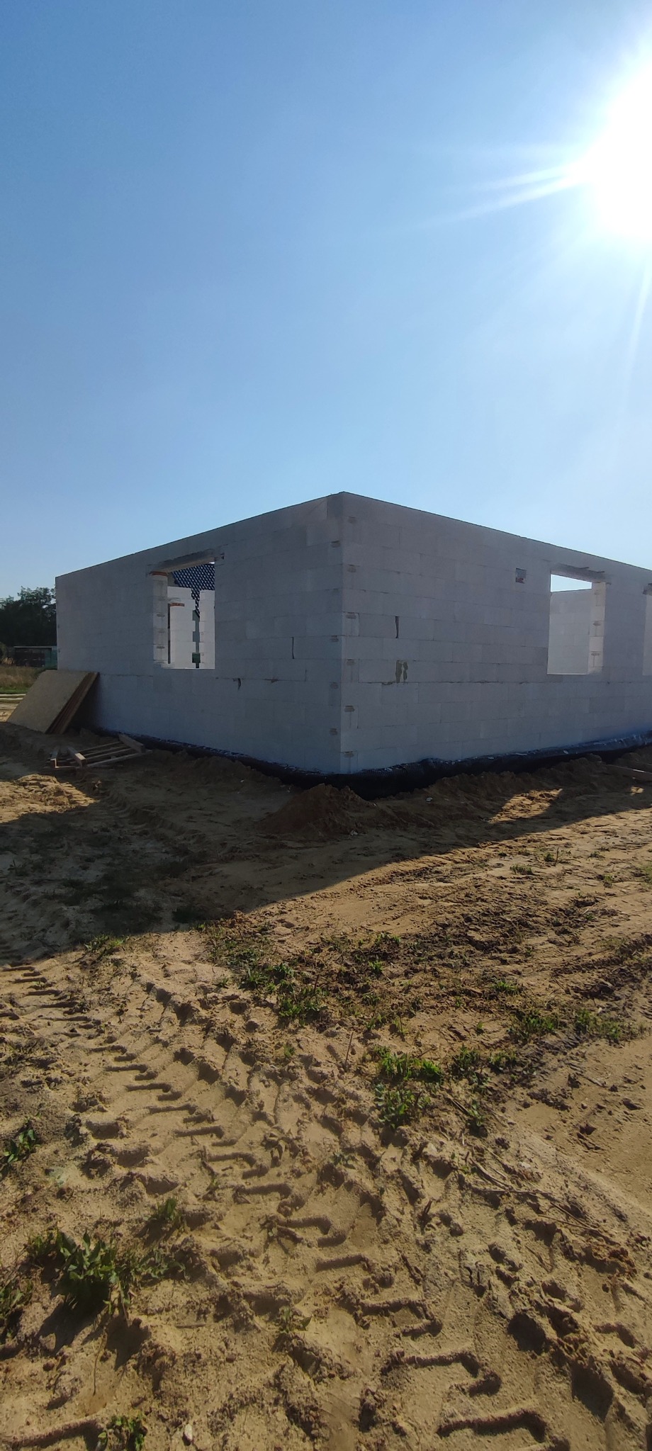 Surowy stan: ściany z betonu komórkowego z otworami okiennymi wzniesione na piaszczystym gruncie, w tle błękitne niebo i zieleń. Widoczne deski i ślady opon.