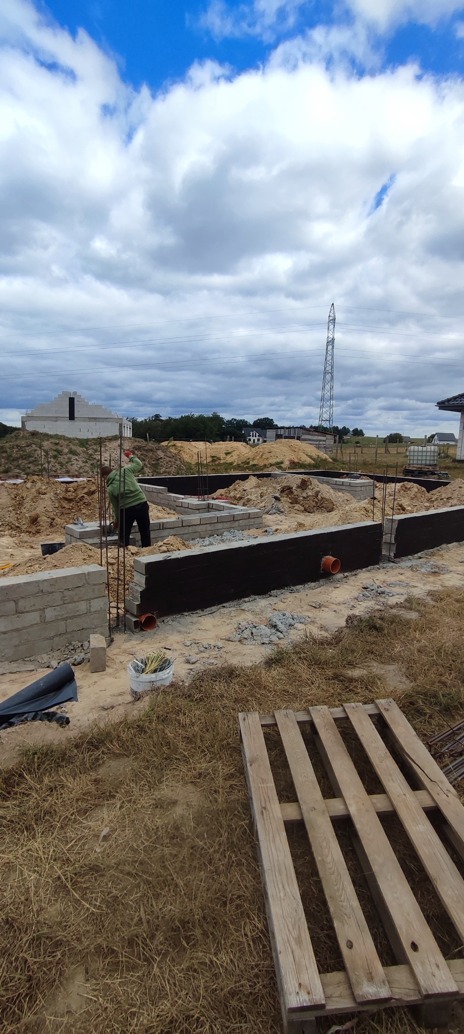 Budowa fundamentów: pracownik w zielonej bluzie układa bloczki, widoczne zbrojenia, izolacja, rury, paleta i ziemia. W tle konstrukcja budynku i słup wysokiego napięcia.