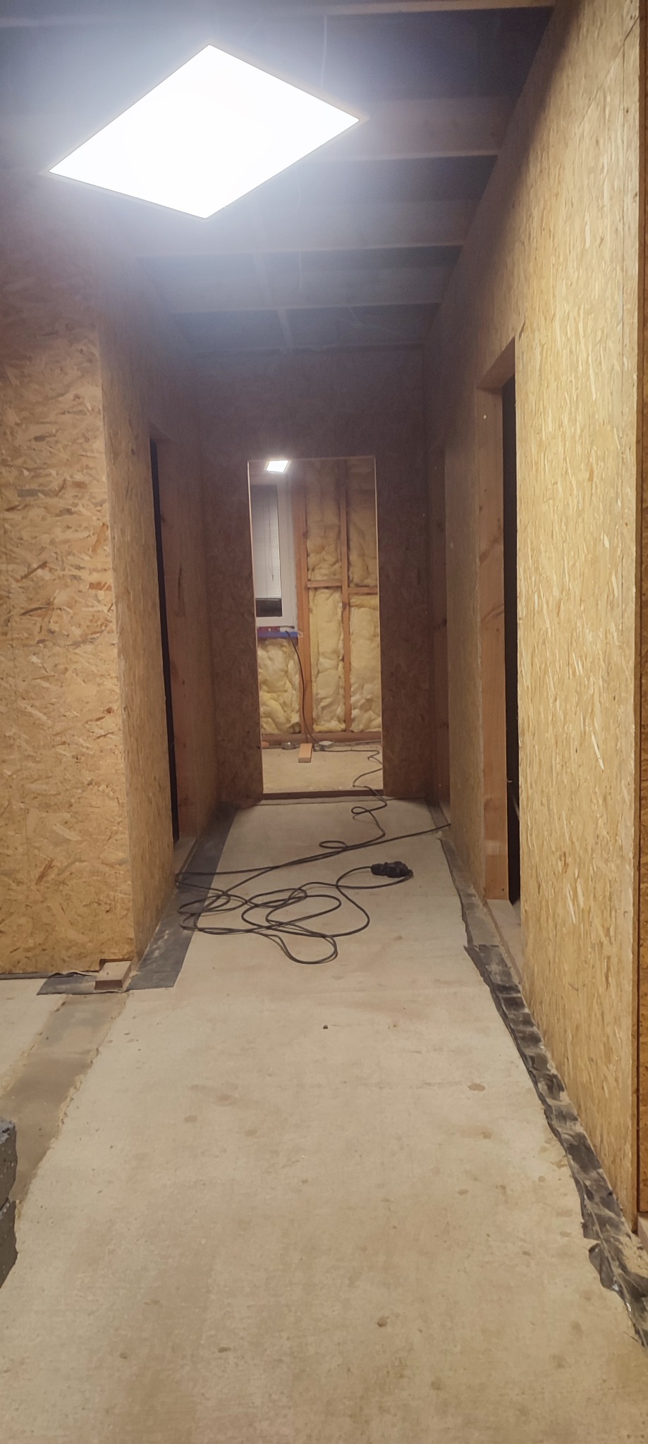 Wnętrze domu w trakcie budowy: ściany z płyt OSB, widoczne ocieplenie w kolejnym pomieszczeniu, surowa podłoga i przewody narzędzi budowlanych.