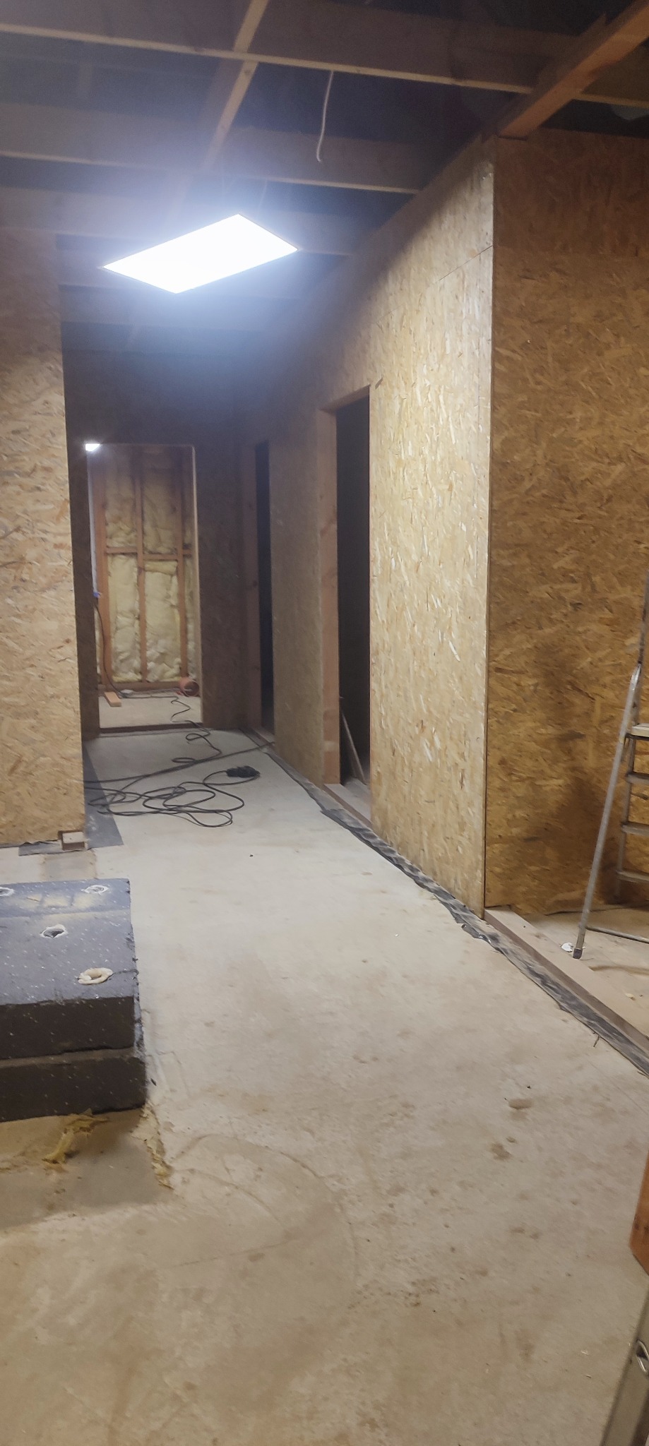 Wnętrze domu w trakcie budowy: surowe ściany z płyty OSB, widoczne instalacje elektryczne i oświetlenie sufitowe. Stan surowy przed wykończeniem.