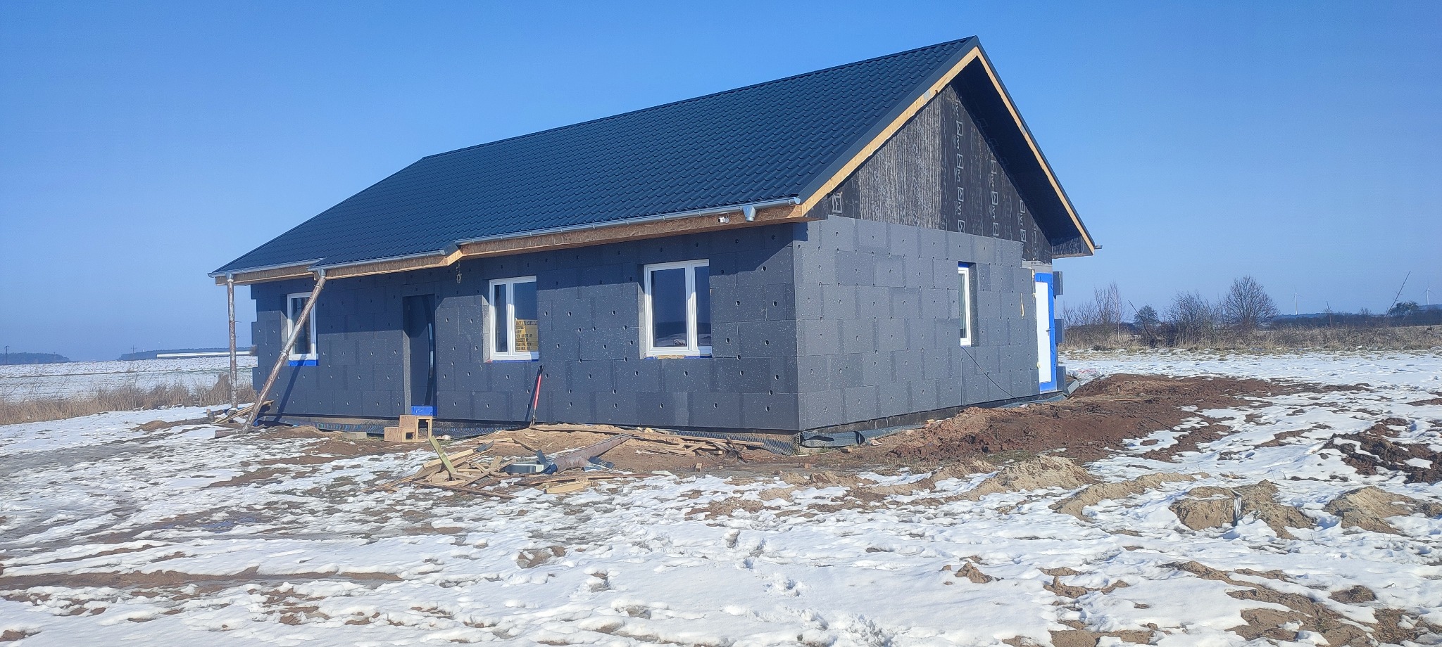 Niedokończony dom z szarej kostki, z oknami i ciemnym dachem, w zimowym krajobrazie z resztkami śniegu na ziemi i błękitnym niebem.