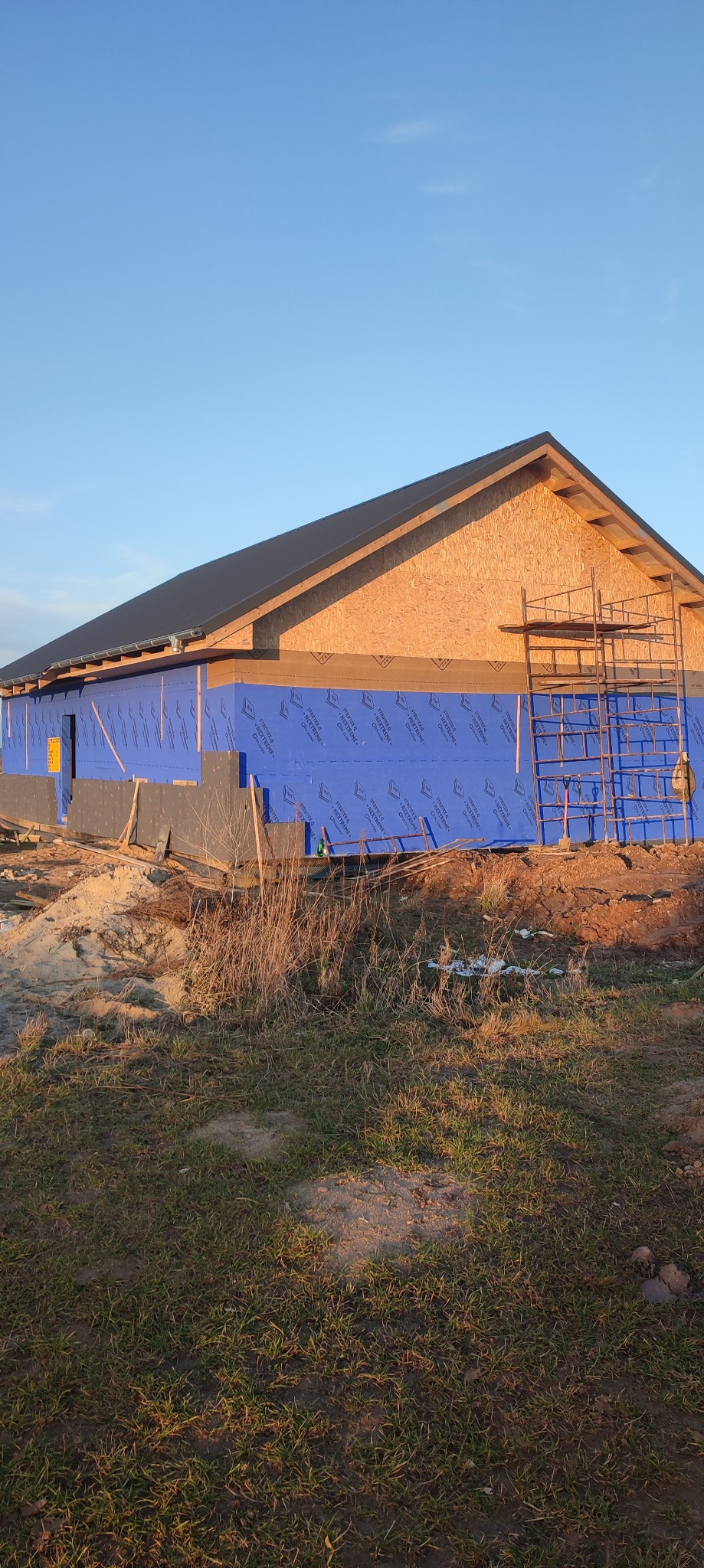 Budowa domu szkieletowego w trakcie: ściany z płyt OSB i niebieskiej membrany, widoczne rusztowanie, dach z czarnej blachy, trawa na pierwszym planie.