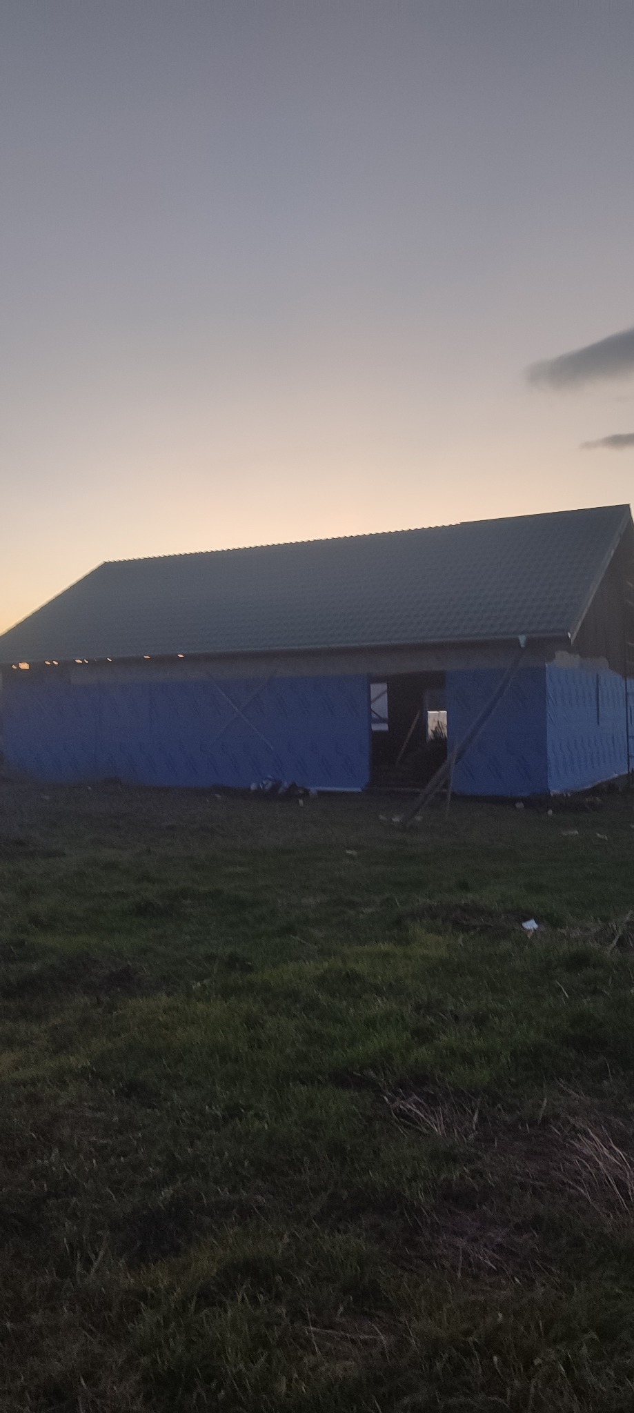 Wznoszony dom z ciemnym dachem i niebieską izolacją na tle zachodzącego słońca. Widoczne zarysy okien i drzwi. Prace budowlane w toku.