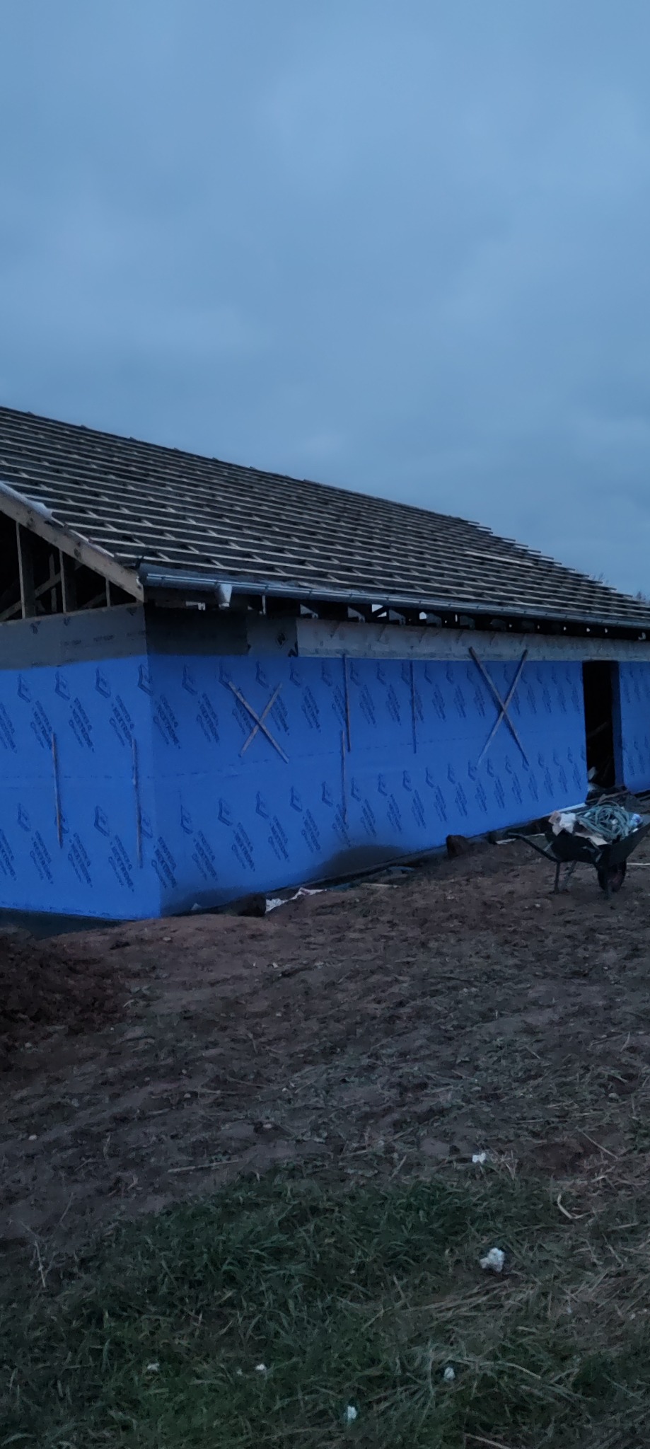 Budowa domu: ściana z niebieską izolacją, widoczny szkielet dachu i taczka. Etap prac budowlanych przy wznoszeniu domu, wieczorne oświetlenie.