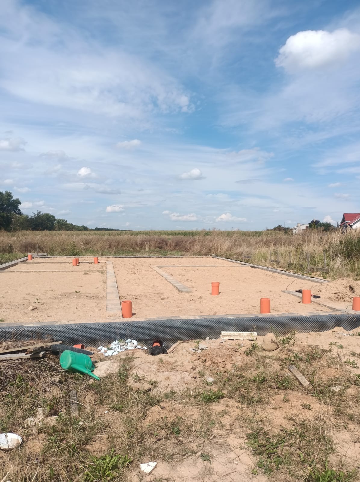 Wykonane fundamenty pod dom z widoczną izolacją, szalunkiem i pionowymi rurami wentylacyjnymi. Tło stanowi łąka i błękitne niebo z chmurami.
