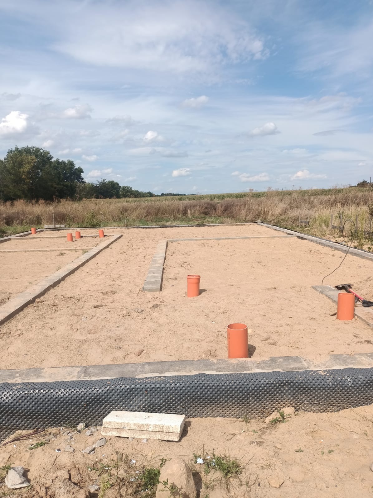 Przygotowany teren pod fundamenty z widoczną izolacją i pionami wentylacyjnymi, na tle łąki i błękitnego nieba z chmurami.