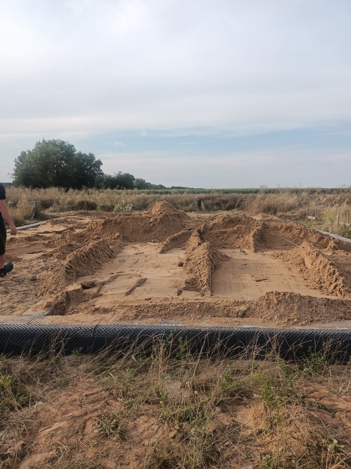 Przygotowanie terenu pod fundamenty domu jednorodzinnego z widocznym zarysem ścian i podkładem z piasku na działce z wysoką trawą.