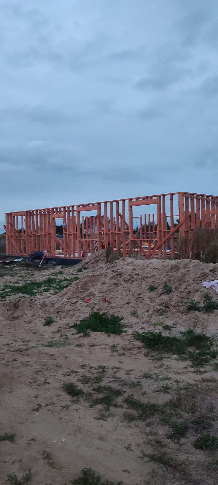 Konstrukcja szkieletowa domu w trakcie budowy, widok na drewniany szkielet z otworami okiennymi na tle pochmurnego nieba i piaszczystego terenu.