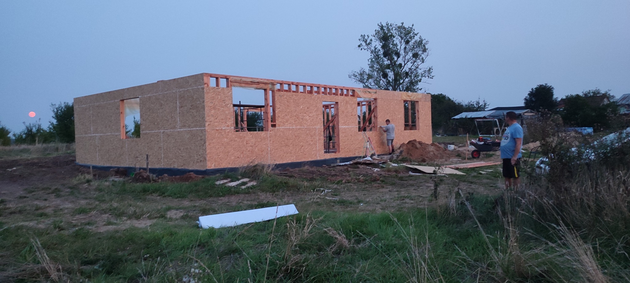 Wznoszenie ścian domu szkieletowego z płyt OSB, widoczny fragment konstrukcji i pracujący mężczyzna. W tle zachodzące słońce nadaje zdjęciu ciepły koloryt.