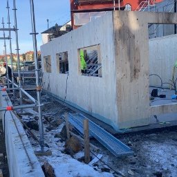 INVEST BYGG Usługi Ogólnobudowlane Jakub Kilian - Wznoszenie drewnianej konstrukcji szkieletowej budynku z widocznymi otworami okiennymi i fragmentami rusztowania, na tle zabudowy miejskiej i błękitnego nieba, w zimowej scenerii z resztkami śniegu.