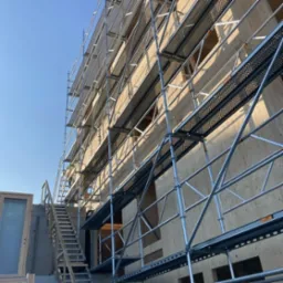 Elewacja budynku w trakcie ocieplania, widoczne rusztowania i płyty izolacyjne z jasnego drewna pod błękitnym niebem.