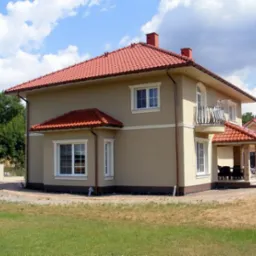 Dwupiętrowy dom z beżową elewacją, czerwoną dachówką i białymi ramami okiennymi, widziany z boku na tle błękitnego nieba z chmurami.