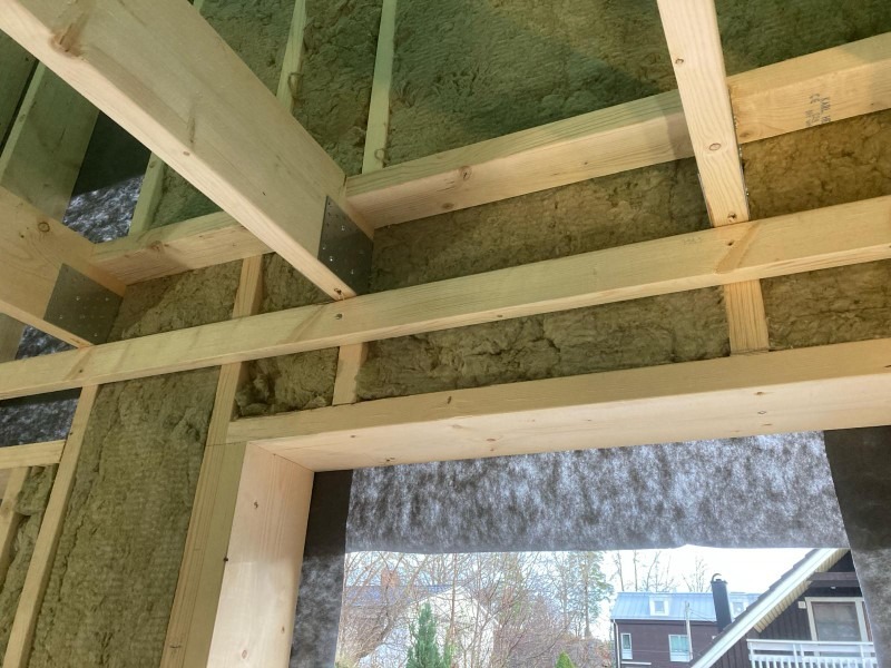 Wnętrze szkieletu budynku z widoczną izolacją z wełny mineralnej między drewnianymi belkami, zabezpieczoną czarną folią paroprzepuszczalną w otworze okiennym.