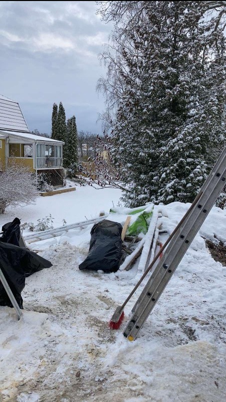 Przygotowania do prac elewacyjnych zimą: drabina oparta o śnieżny grunt, worki na odpady, deski i szczotka przed żółtym domem z przeszkloną werandą.