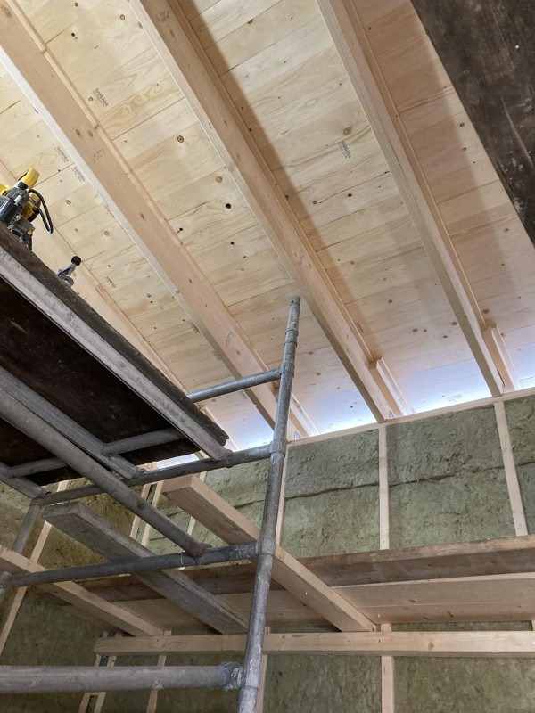 Wnętrze w trakcie prac: widoczne ocieplone ściany wełną mineralną, drewniana konstrukcja dachu z deskowaniem, rusztowanie i narzędzia na górnej platformie.
