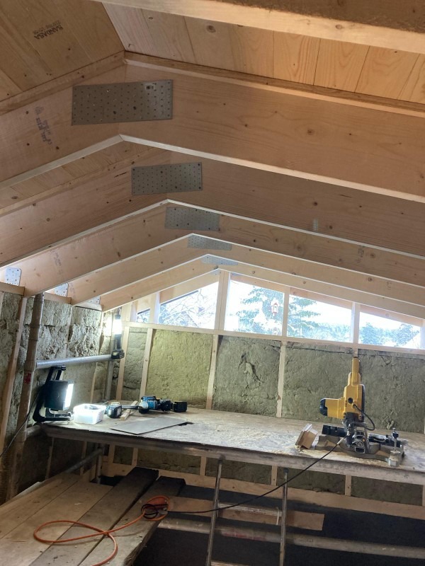 Wnętrze budynku w trakcie izolacji, widoczne wełna mineralna na ścianach, drewniana konstrukcja dachu wzmocniona metalowymi płytkami, narzędzia pracy na prowizorycznym stole warsztatowym, okno...