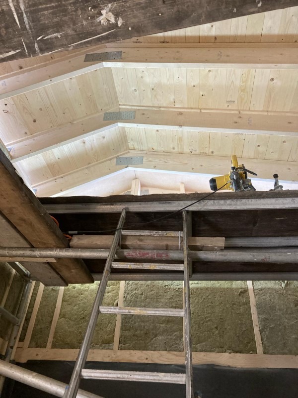 Poddasze w trakcie prac wykończeniowych: widoczne drewniane panele na suficie, izolacja z wełny mineralnej w ścianach, rusztowanie i drabina, narzędzia do montażu paneli na górnym poziomie.