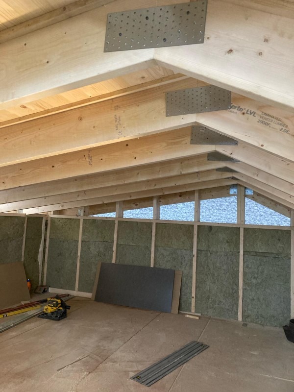 Wnętrze poddasza w trakcie izolacji: drewniane belki stropowe z metalowymi wzmocnieniami, ściany wyłożone wełną mineralną, widoczne narzędzia pracy i płyty izolacyjne.