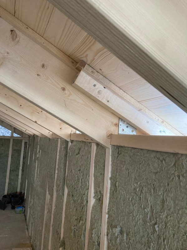 Wnętrze poddasza w trakcie prac: drewniana konstrukcja dachu z widocznym ociepleniem ścian wełną mineralną między słupkami. Metalowe łączniki wzmacniają konstrukcję.