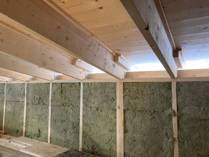 Wnętrze w trakcie prac izolacyjnych: drewniana konstrukcja szkieletowa wypełniona wełną mineralną, widoczne elementy mocujące i surowy sufit z desek.