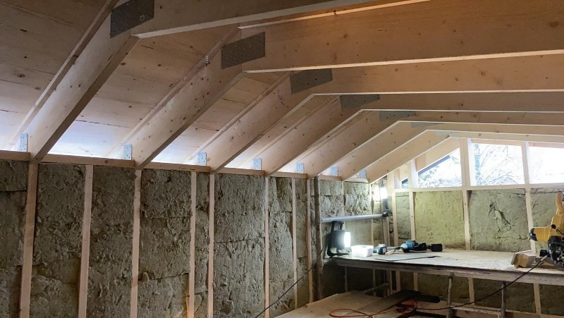 Wnętrze poddasza w trakcie prac: izolacja ścian wełną mineralną pomiędzy drewnianymi słupkami, widoczne elementy konstrukcji dachu z metalowymi łącznikami, okno w tle, narzędzia na prowizorycznym...