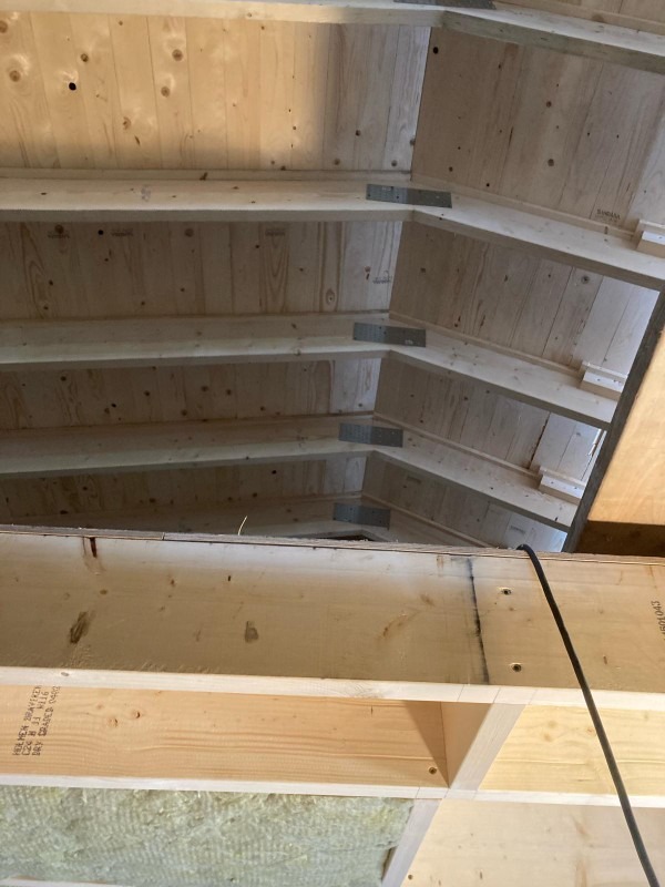 Surowe drewniane krokwie dachowe połączone metalowymi łącznikami, widoczne ocieplenie wełną mineralną, czarny przewód elektryczny w tle.