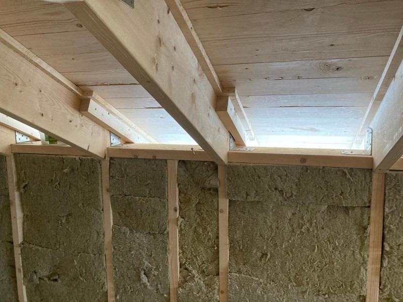 Wnętrze budynku w trakcie prac: drewniana konstrukcja stropu z widocznymi metalowymi łącznikami i wełna mineralna między słupkami ściany.