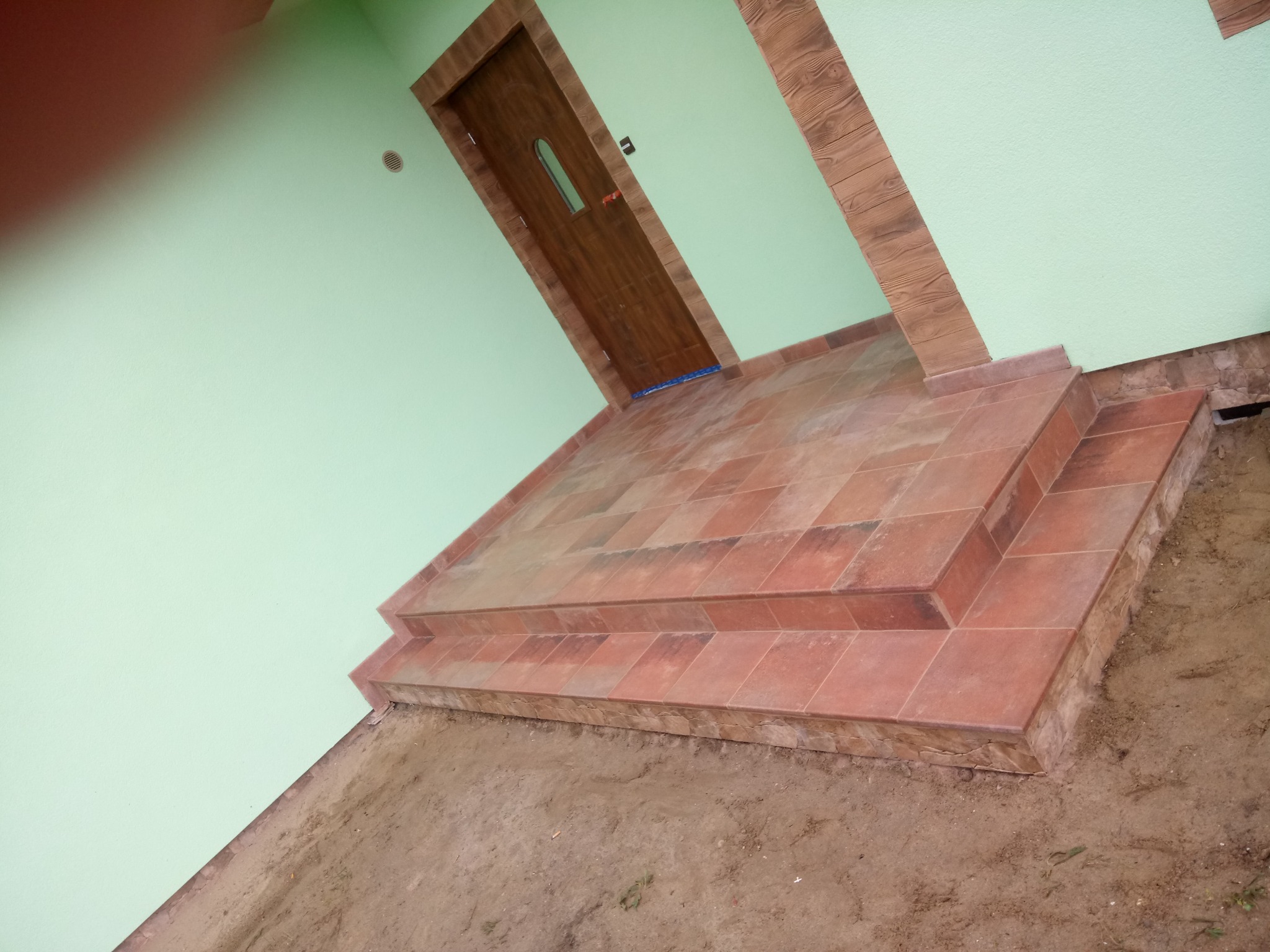 Wykończone kafelkami schody wejściowe do domu z zieloną elewacją i brązowymi drzwiami, widok z dołu na zewnątrz budynku, częściowo widoczny niezabezpieczony grunt.