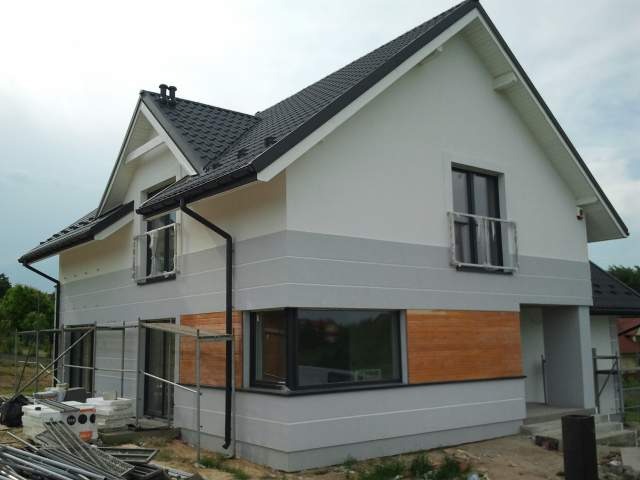 Dwukondygnacyjny dom z szarą i białą elewacją, częściowo wykończoną drewnem, widoczne rusztowania i materiały budowlane, dach pokryty czarną dachówką