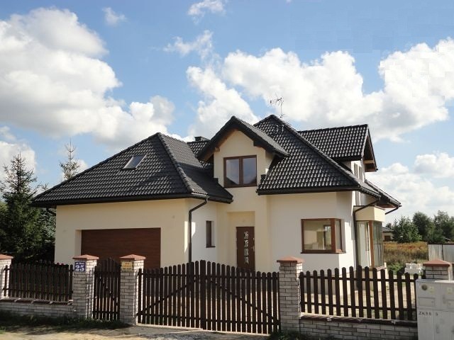 Jasny, dwukondygnacyjny dom jednorodzinny z ciemną dachówką, widoczny zza drewnianego ogrodzenia z ceglanymi słupkami, błękitne niebo z obłokami w tle.