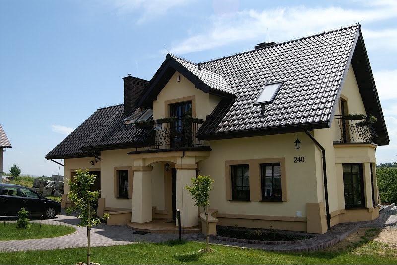 Elewacja domu jednorodzinnego w jasnym odcieniu z ciemną dachówką, balkonem z kutą balustradą i numerem domu 240, otoczona zielenią i brukowanym chodnikiem.