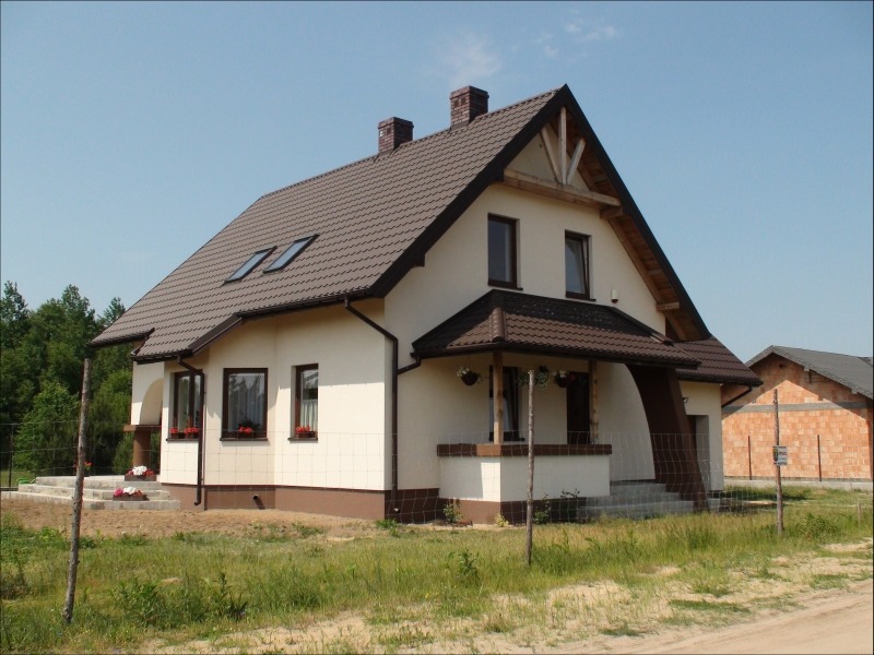 Dwukondygnacyjny dom z brązowym dachem, jasną elewacją i kwiatami w oknach, widoczny fragment budynku w trakcie budowy w tle.