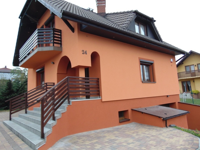 Dwukondygnacyjny dom jednorodzinny z balkonem, pomalowany na pomarańczowo, z brązowymi elementami wykończeniowymi, numer '24' nad wejściem, widok z zewnątrz.