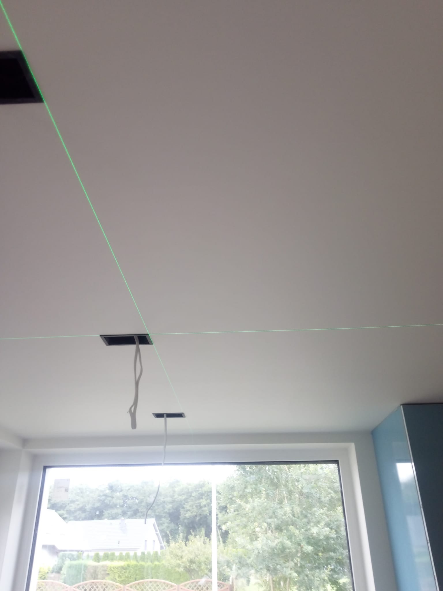 Biały sufit z dwoma otworami na oświetlenie, z których zwisają kable, wyznaczony laserowo krzyż linii poziomych i pionowych, okno w tle.
