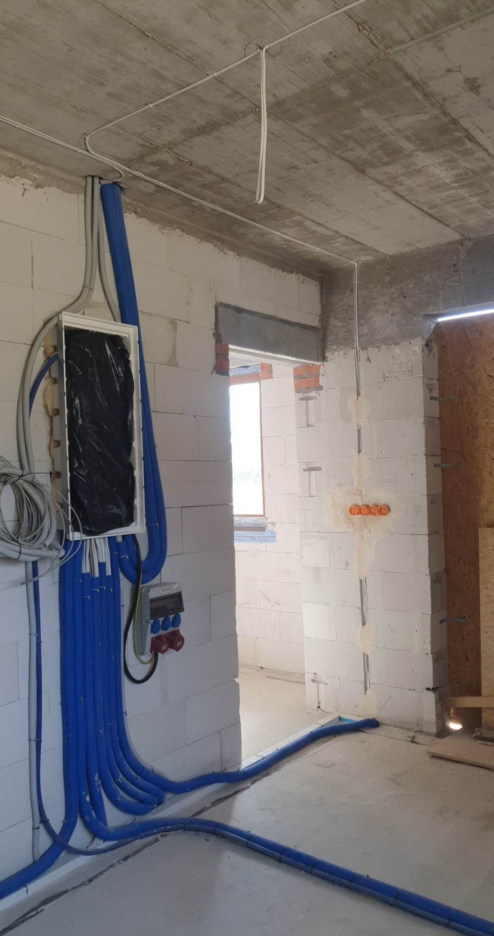 Niedokończona instalacja elektryczna w surowym wnętrzu: niebieskie rury karbowane prowadzą do skrzynki zabezpieczającej, widoczne przewody i puszki elektryczne w ścianach z betonu komórkowego.